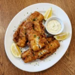 Crispy beer battered fish on a white oval ceramic dinner plate with lemon wedges and tartar sauce