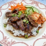 Korean Beef Bulgogi Bowl with browned beef rice cucumbers carrots kimchi and sesame seeds on a white plate