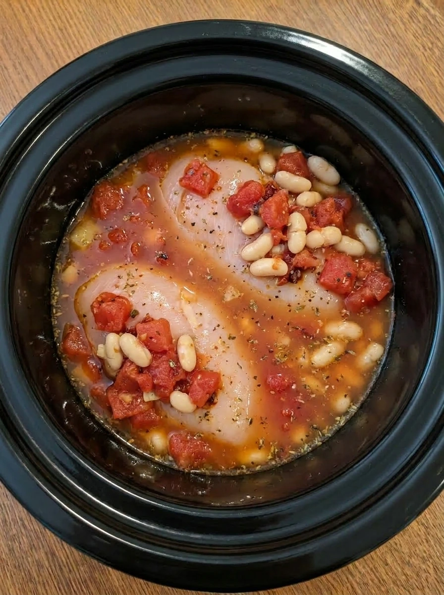 Broth, tomatoes, beans, and seasonings added to chicken in a slow cooker.